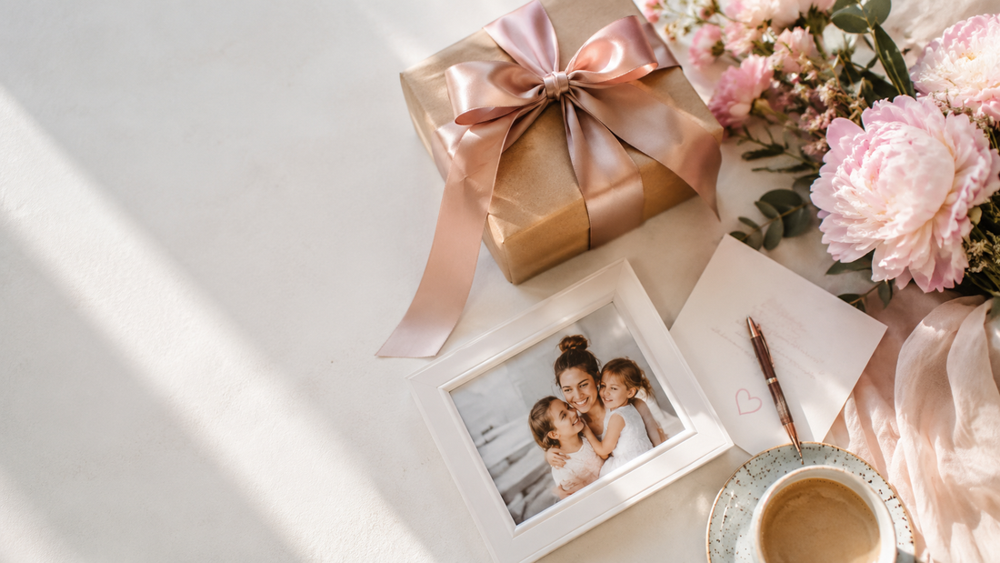 מתנה לאמא — flat lay עם קופסת מתנה זהובה, מסגרת תמונה של אם וילדות ופרחים